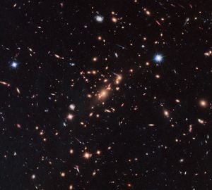 A bunch of bright dots, spheres, and spirals of light (mostly red-orange, with some blue and white) against the black backdrop of space, with a clump near the center that have the circular lensing effect. A bright blue foreground star in the upper-right.